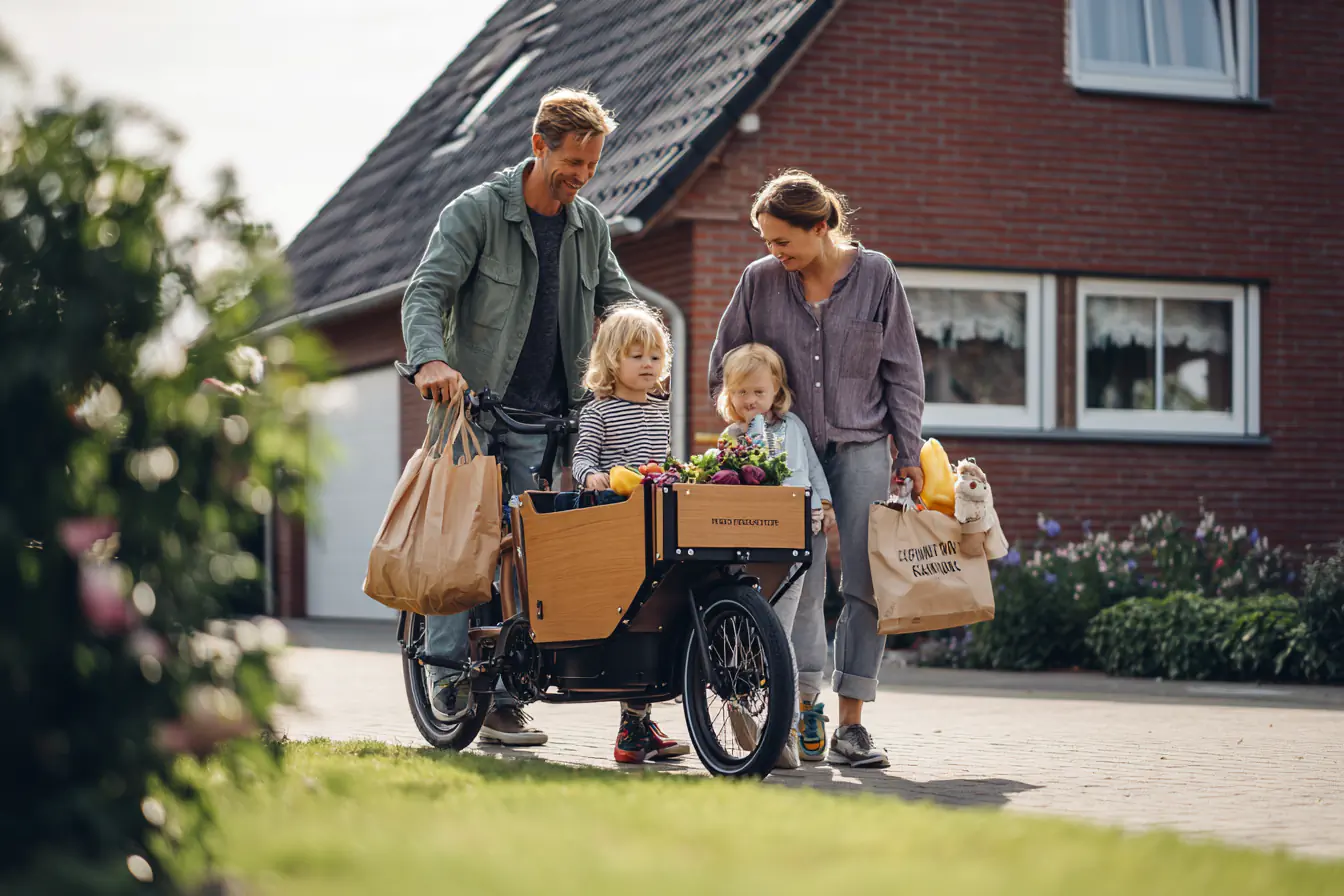 Glückliche Familie mit zwei Kindern und Einkäufen auf einem Lastenrad vor einem Haus.